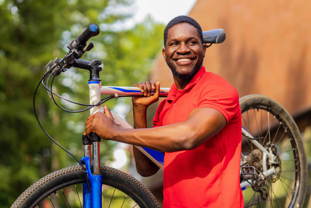 afro american man in red t-shirt walking in park with a bicycle. - Photo, Image