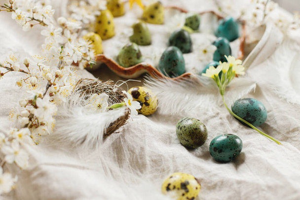 Happy Easter! Stylish easter eggs and blooming spring flowers on rustic table. Natural painted quail eggs in tray, feathers and cherry blossoms on linen fabric. Rustic easter still life - Foto, afbeelding