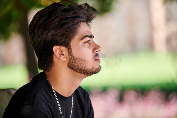 Homme indien attrayant fumeur exhale portrait de fumée de cigarette en t-shirt noir et chaîne de cou en argent dans le parc public, hindou fumeur masculin portrait rapproché. Beau portrait d'homme indien aux cheveux épais - Photo, image