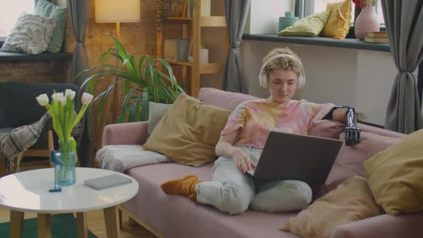 Chica con el brazo protésico sentado en el sofá en la sala de estar y viendo la película en línea en el ordenador portátil con auriculares inalámbricos - Imágenes, Vídeo