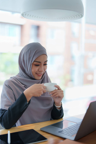 Красива мусульманка з усмішкою на обличчі п "є каву і комп" ютер.. - Фото, зображення