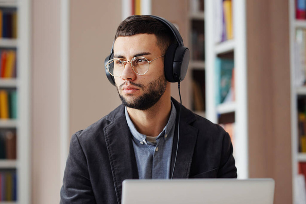 Bearded man with eyeglasses sitting in library, working on laptop and listening to audio course or podcast with headphones, making pause and looking away. Student studying, preparing assignment - Photo, image