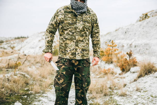 Soldier is standing in the camouflage uniform and checkered keffiyeh shemagh bandana. Man with knife is outdoors in the abandoned deserted place. - Foto, Bild