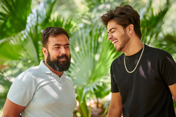 Two funny indian men laughing and speak Hindi in public park green palm leaves background, true male friendship. Two amusing indian guys laughing out loud on outdoor walking in park - Φωτογραφία, εικόνα