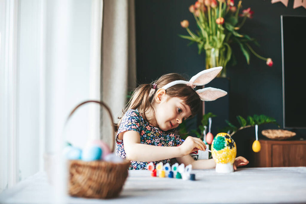 Little girl with bunny ears painting gypsum Easter egg with paintbrush. Easter celebration concept. - 写真・画像