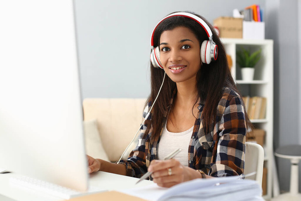 African female student wears headphones studying alone at home office desk. Mixed race woman listening to audio podcast and learning english language concept and taking notes concept - Fotoğraf, Görsel