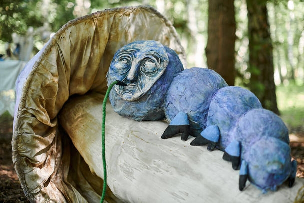 Гусеница для курения кальян от объекта искусства "Страна чудес Алисы" на выставке в зеленом лесу, атмосферный сюрреализм. Курящая гусеница лежит на большом грибе, объекте сказочного искусства - Фото, изображение