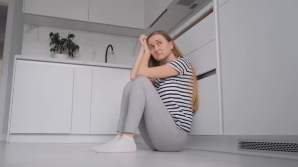 Μια κουρασμένη γυναίκα ευρωπαϊκής εμφάνισης κάθεται στο πάτωμα της κουζίνας και ξεκουράζεται. Μια γυναίκα σε κατάθλιψη και απελπισία - Πλάνα, βίντεο