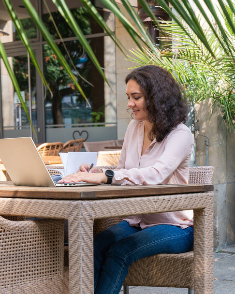 Una joven freelancer de negocios se sienta en una mesa en un café de la calle y trabaja en una computadora portátil y toma notas en un cuaderno con un lápiz. - Foto, imagen