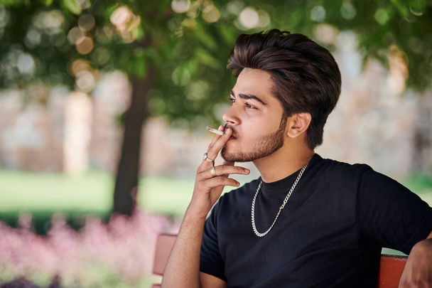 Jonge indiaan roker portret in zwart t-shirt en zilveren halsketting zittend op bank in openbaar park, hindoe man roken close-up portret. Knappe indiaanse man portret met dik haar in stadspark - Foto, afbeelding