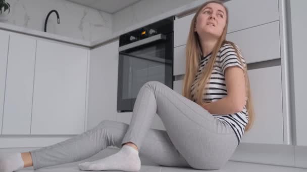 Nainen, jolla on vatsakipuja kuukautisten aikana, istuu lattialla keskellä keittiötä. Nuori nainen kärsii kuukautisensa aikana - Materiaali, video