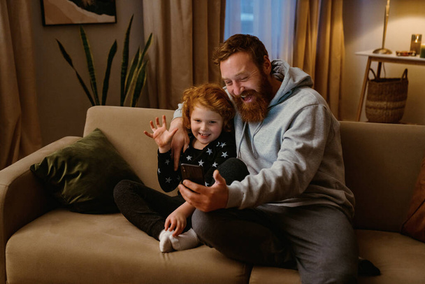 Excited father and son having video call via mobile phone while sitting on sofa in cozy living room - Fotografie, Obrázek