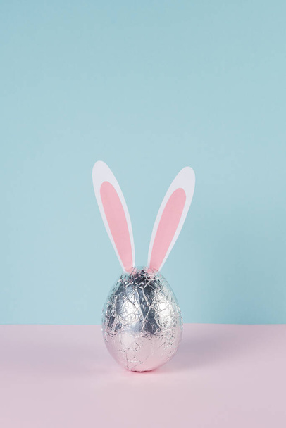Easter egg wrapped in a silver foil with bunny or rabbit ears. Minimal pastel pink and blue Easter holiday concept. - Valokuva, kuva