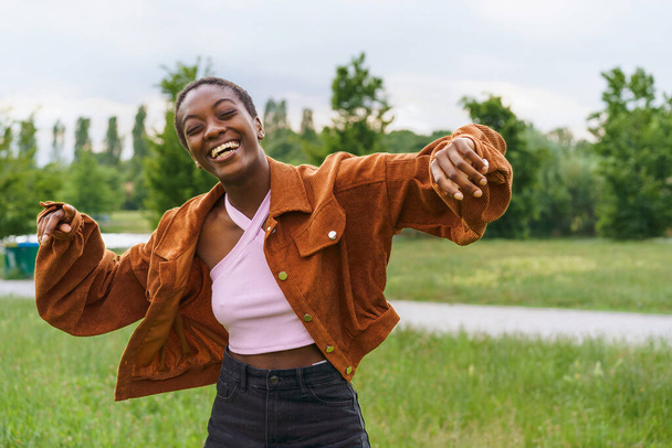 Μια νεαρή Αφρο-Αμερικανίδα με κοντά μαλλιά φαίνεται να χορεύει και να γελάει σε δημόσιο πάρκο την άνοιξη - ένα μαύρο κορίτσι που φοράει καθημερινά ρούχα, αποπνέοντας χαρά, ζωντάνια και νεανικότητα - Φωτογραφία, εικόνα