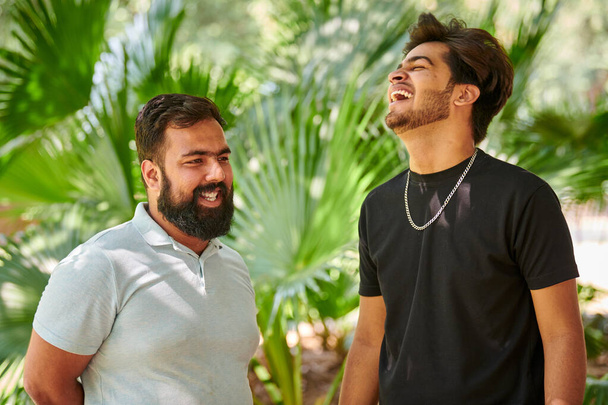 Dwóch zabawnych Indian śmiejących się i mówiących po hindusku w parku publicznym zielona palma pozostawia tło, prawdziwą męską przyjaźń. Dwóch zabawnych Indian śmiejących się na głos na świeżym powietrzu spacerujących w parku - Zdjęcie, obraz
