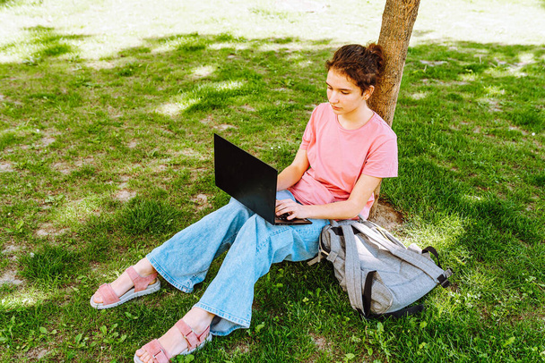 Adolescente estudiante, mujer joven, sentada en el césped en el parque, usando jeans, camiseta rosa, con computadora portátil, estudiando en línea, haciendo tareas, aprendiendo, redes sociales. Retrato de adolescente que estudia en línea en el parque en verano - Foto, imagen