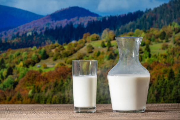 Fresh white milk in a jug and glass on wooden table with the autumn Carpathian mountains background, close up. Dairy product concept - Valokuva, kuva