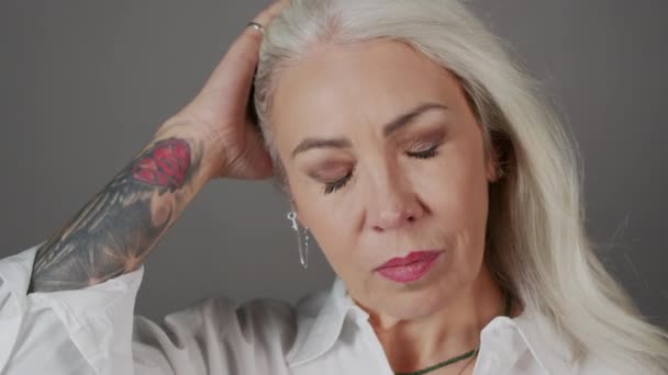Closeup portrait of attractive mature woman with arm tattoo and blowing long grey hair looking at camera at dark grey studio background - Footage, Video
