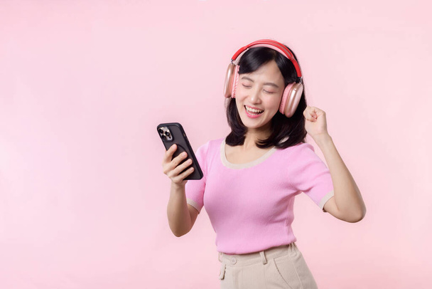 Portrait junge fröhliche asiatische Frau genießen Audio-Hören von Smartphone-Musik-Anwendung gegen rosa. Glücklich lächelnde Frau mit Kopfhörer. Sound, Freizeit, Lifestyle, Technologiekonzept - Foto, Bild