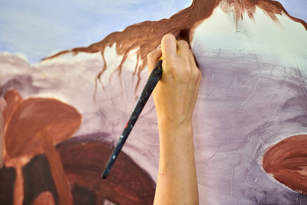 Girl artist hand holds paint brush and draws abstract surreal landscape on white canvas at outdoor art painting festival, paintings art picture process. Woman artist paints atmospheric surreal picture - 写真・画像
