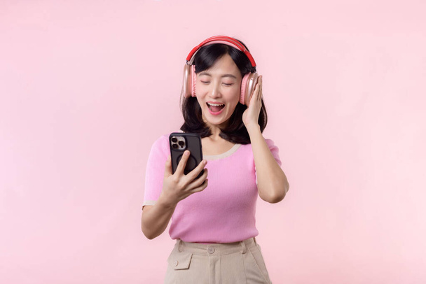 Portrait young cheerful asian woman enjoy listening audio by smartphone music application against pink. Happy smiling female person with headphone. Sound, leisure, lifestyle, technology concept - Zdjęcie, obraz