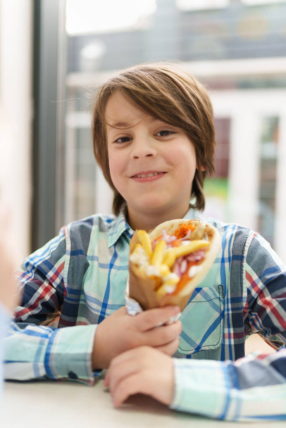 Happy little boy holding gyros wrap in hands. Portrait of cheerful white kid eating Greek fast food in a cafe - Foto, immagini