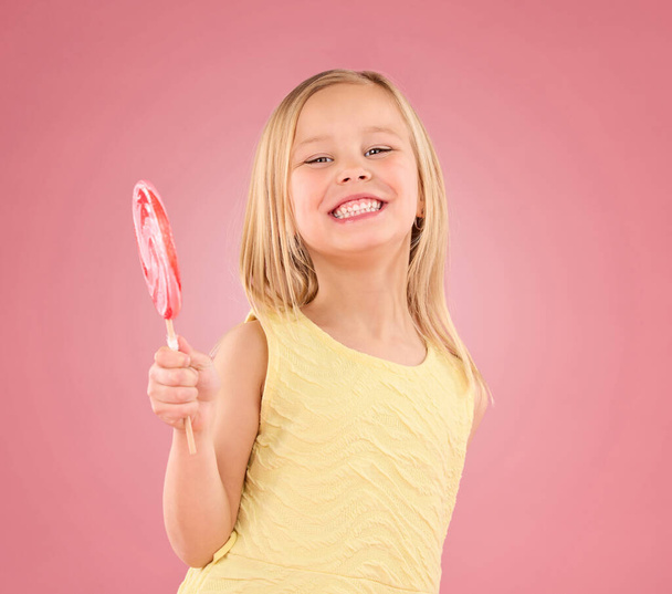 Candy, úsměv a lízátko s portrétem dívky ve studiu pro cukr, party a karnevalové jídlo izolované na růžovém pozadí. Roztomilé, pozitivní a mládež s dítětem a jíst svačinu pro hravé a lahůdky. - Fotografie, Obrázek