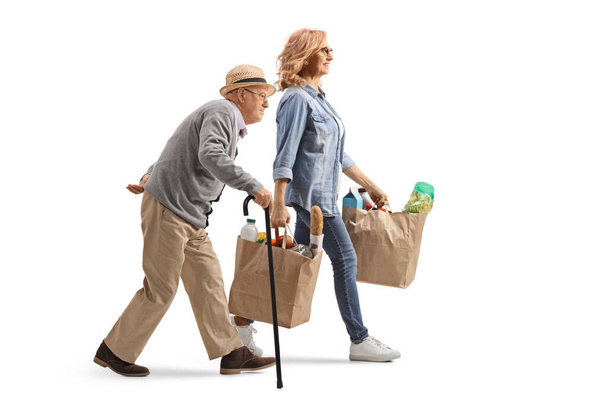 Täyspitkä profiilikuva vanhuksesta kävelemässä kävelykepin kanssa ja naisesta kantamassa valkoisella pohjalla olevia ruokakasseja - Valokuva, kuva
