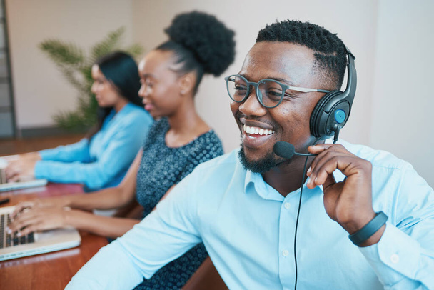 Ένας ελκυστικός μαύρος άντρας μιλάει στον πελάτη του σε τηλεχειριζόμενο ακουστικό, χαμογελώντας. Υψηλής ποιότητας φωτογραφία - Φωτογραφία, εικόνα
