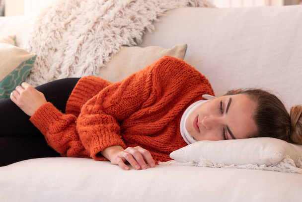 Κουρασμένη νεαρή γυναίκα με πορτοκαλί ζεστά ρούχα με ορθοπεδικό γιακά ξαπλωμένη στον καναπέ για να ξεκουραστεί μετά από τραυματισμό του τραχήλου της μήτρας στο σαλόνι στο σπίτι - Φωτογραφία, εικόνα
