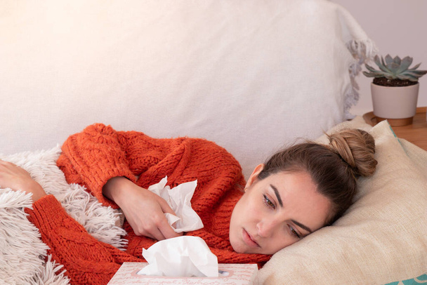 Femme malade avec allergies de saison reposant avec nez rouge sur le canapé sous couverture floue dans le salon moderne avec des mouchoirs en papier et regardant loin - Photo, image