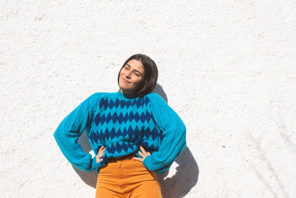 Smiling young latin girl in colorful clothes, saturated blue argyle sweater and orange pants, white stucco wall background outside, light sunny day. Copy space on one side - Photo, Image
