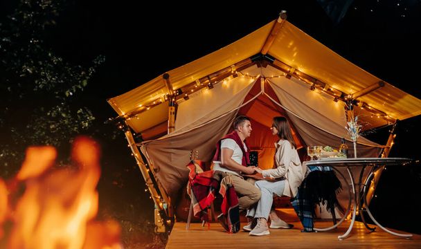 Happy lovely couple relaxing in glamping on summer evening and drinking wine near cozy bonfire. Luxury camping tent for outdoor recreation and recreation. Lifestyle concept - Fotó, kép