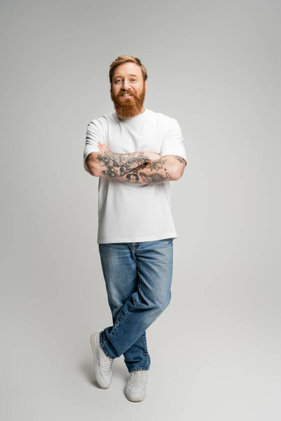 Full length of smiling tattooed man crossing arms on grey background - 写真・画像