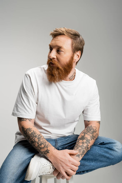 Tattooed man in jeans and t-shirt sitting on chair isolated on grey  - Photo, image