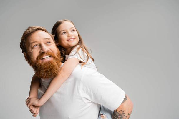 Cheerful child looking away while piggybacking on bearded father isolated on grey  - Fotografie, Obrázek