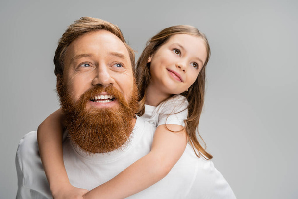 Preteen kid looking away while piggybacking on smiling bearded dad isolated on grey  - Fotografie, Obrázek