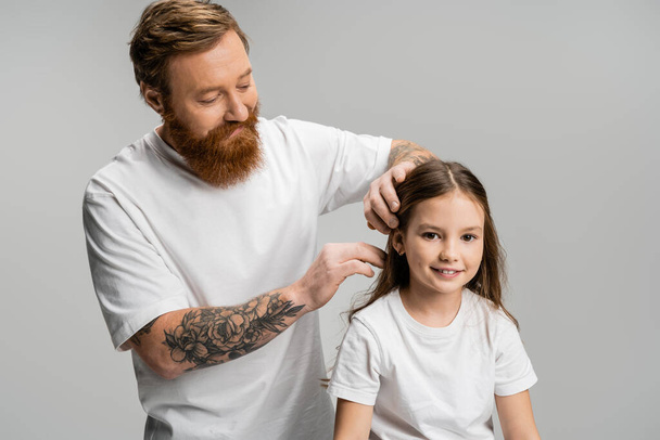Bearded father adjusting hair of smiling preteen daughter isolated on grey  - Fotografie, Obrázek