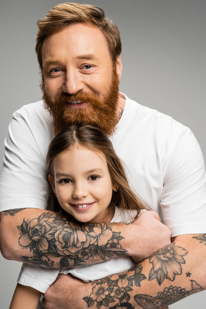 Portrait of tattooed father looking at camera while hugging smiling preteen girl isolated on grey  - Фото, зображення