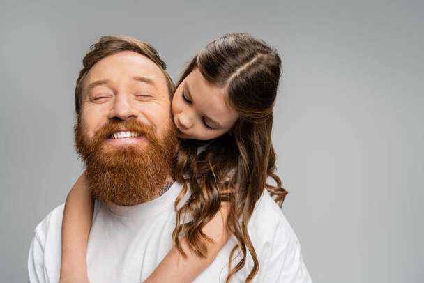 Preadolescente chica besándose y abrazando sonriente barbudo papá aislado en gris  - Foto, imagen