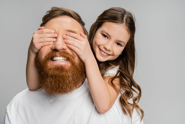 Cheerful preteen girl covering eyes of happy father with beard isolated on grey  - Zdjęcie, obraz