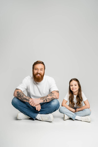Positive tattooed father and preteen kid looking at camera while sitting on grey background - 写真・画像