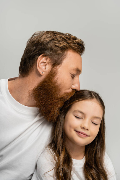Bearded dad kissing head of smiling preteen daughter with closed eyes isolated on grey   - Foto, afbeelding