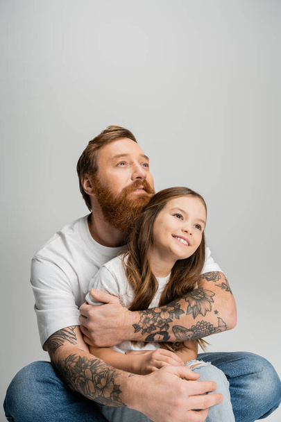 Τατουάζ άντρας αγκαλιάζει ανέμελη κόρη και κοιτάζει μακριά απομονωμένος στο γκρι   - Φωτογραφία, εικόνα
