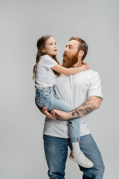 Tattooed man holding preteen daughter and talking isolated on grey   - Foto, afbeelding