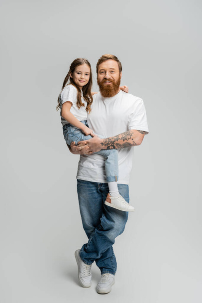Full length of positive father in t-shirt and jeans holding daughter on grey background - Photo, Image