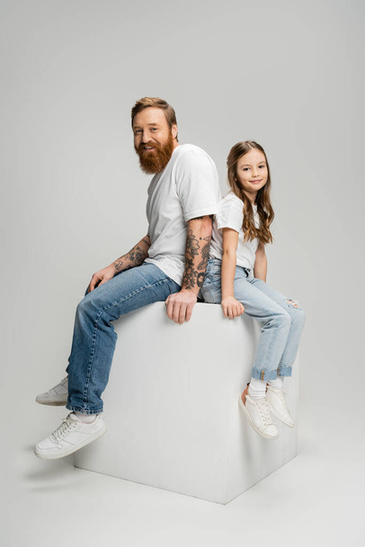 Cheerful tattooed man sitting on cube near daughter on grey background - Photo, image