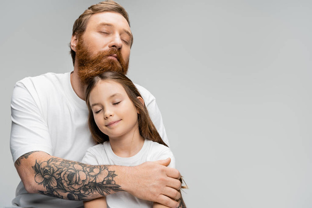 Tattooed man with closed eyes hugging smiling preteen daughter isolated on grey   - Photo, Image