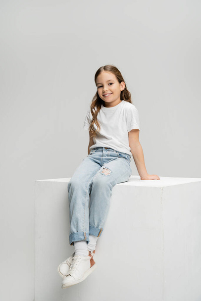 Cheerful kid in t-shirt and jeans sitting on cube isolated on grey   - Photo, Image