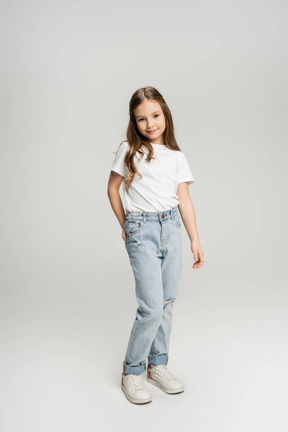 Full length of preteen girl in jeans and t-shirt posing and smiling on grey background  - Фото, изображение
